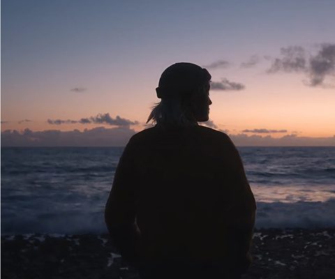 Silhouette of a person standing on a shoreline at sunset, looking out over the ocean with soft light reflecting on the waves.