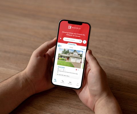 Photo of a person holding a smartphone displaying the REALTOR.ca app, with a property listing visible on the screen.