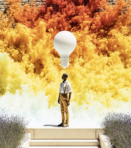 Surreal artwork of a person standing before a dramatic cloud of yellow, orange, and red smoke, with a large white light bulb floating above their head—symbolizing strategy, ideas, and creative vision.